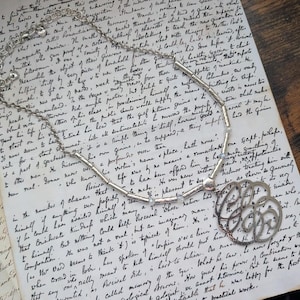 K&ouml;nnte beinhalten: Silberfarbene Halskette mit einem Anh&auml;nger mit filigranem Design. Die Kette liegt auf einem alten, handgeschriebenen Dokument. Die Kette hat kleine, klare Perlen. Das Design wirkt klassisch und elegant.