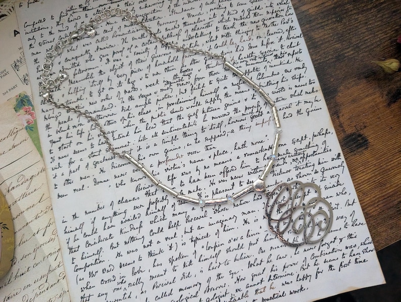 K&ouml;nnte beinhalten: Silberfarbene Halskette mit einem Anh&auml;nger mit filigranem Design. Die Kette liegt auf einem alten, handgeschriebenen Dokument. Die Kette hat kleine, klare Perlen. Das Design wirkt klassisch und elegant.