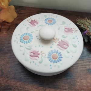 Puede incluir: Caja de cerámica blanca con tapa y diseño floral. La tapa presenta flores rosas y azules con hojas verdes sobre un fondo blanco. La caja tiene un pomo blanco en la parte superior.
