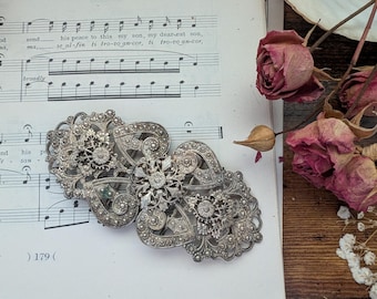 May include: An ornate, silver-toned vintage clasp with intricate filigree and sparkling accents. The clasp is set against a backdrop of aged sheet music and dried roses, evoking a sense of history and elegance. The clasp is approximately 4 inches wide.