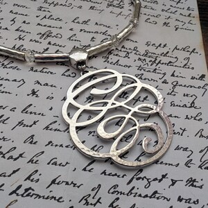 K&ouml;nnte beinhalten: Silberfarbene Monogramm-Halskette mit geh&auml;mmerten Texturen, mit einem gro&szlig;en, verzierten Initialen-Anh&auml;nger. Die Halskette wird auf einem Vintage-Papierhintergrund mit handschriftlichem Skript pr&auml;sentiert, was eine klassische und elegante &Auml;sthetik schafft.