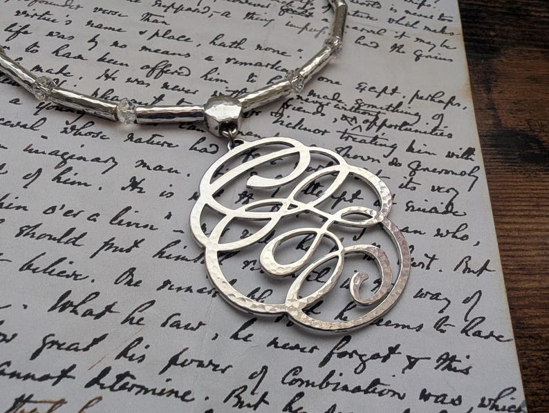 K&ouml;nnte beinhalten: Silberfarbene Monogramm-Halskette mit geh&auml;mmerten Texturen, mit einem gro&szlig;en, verzierten Initialen-Anh&auml;nger. Die Halskette wird auf einem Vintage-Papierhintergrund mit handschriftlichem Skript pr&auml;sentiert, was eine klassische und elegante &Auml;sthetik schafft.