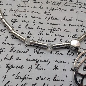 K&ouml;nnte beinhalten: Eine silberne Halskette mit geh&auml;mmerten Texturen und klaren Kristallakzenten. Die Halskette hat einen Anh&auml;nger mit einem Schleifendesign. Der Hintergrund ist ein handgeschriebenes Dokument.