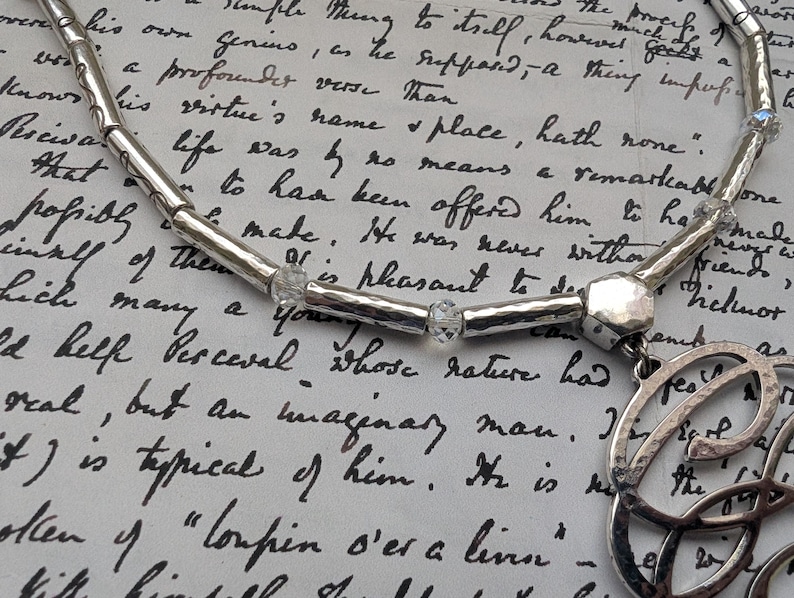 K&ouml;nnte beinhalten: Eine silberne Halskette mit geh&auml;mmerten Texturen und klaren Kristallakzenten. Die Halskette hat einen Anh&auml;nger mit einem Schleifendesign. Der Hintergrund ist ein handgeschriebenes Dokument.