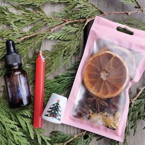 Może przedstawiać: Różowa torebka z przezroczystym okienkiem, w którym znajdują się suszone plasterki pomarańczy, anyż gwiazdkowy i inne przyprawy. W zestawie znajduje się również mała butelka olejku eterycznego i czerwona świeca.