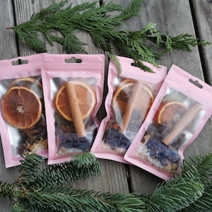 Puede incluir: Cuatro bolsas rosas con ventanas transparentes que contienen rodajas de naranja secas, palitos de canela y otras especias. Las bolsas están etiquetadas con "Spice Winter Pot".