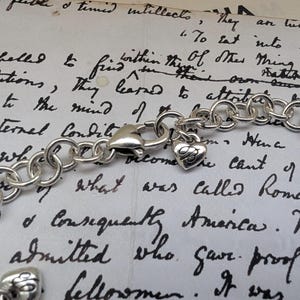 K&ouml;nnte beinhalten: Ein silberfarbenes Charm-Armband mit einem herzf&ouml;rmigen Anh&auml;nger und einem Karabinerverschluss. Das Armband besteht aus miteinander verbundenen, kreisf&ouml;rmigen Gliedern. Der Herzanh&auml;nger hat eine eingravierte Initiale. Der Hintergrund ist ein handgeschriebenes Dokument.