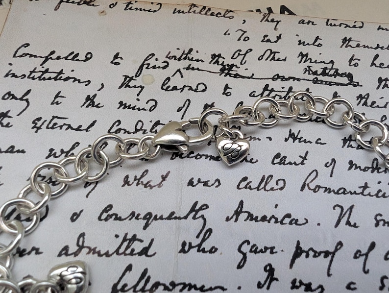 K&ouml;nnte beinhalten: Ein silberfarbenes Charm-Armband mit einem herzf&ouml;rmigen Anh&auml;nger und einem Karabinerverschluss. Das Armband besteht aus miteinander verbundenen, kreisf&ouml;rmigen Gliedern. Der Herzanh&auml;nger hat eine eingravierte Initiale. Der Hintergrund ist ein handgeschriebenes Dokument.