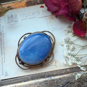 May include: A vintage brooch with a large, oval, light blue stone set in a silver tone metal frame. The brooch is on a postcard with the text "TUCK'S POSTAL CARTE POSTALE".