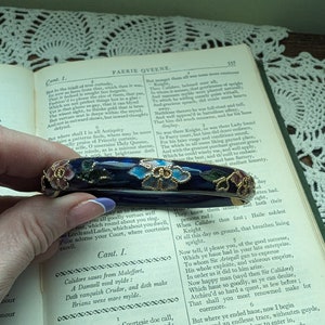 May include: A blue enamel bangle bracelet with floral designs in pink, blue, and gold. The bracelet is on a book with text.