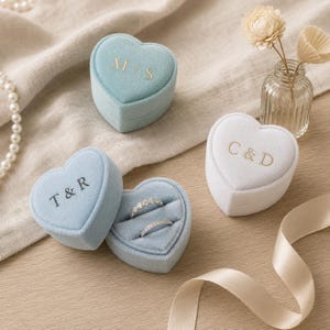 Peut inclure: Trois boîtes à bagues en velours en forme de cœur, bleu clair et blanc. Une boîte bleue est ouverte, révélant deux bagues en argent. Les boîtes portent des initiales gaufrées dorées ou noires : M/S, T & R et C & D, avec un vase en verre et un ruban.