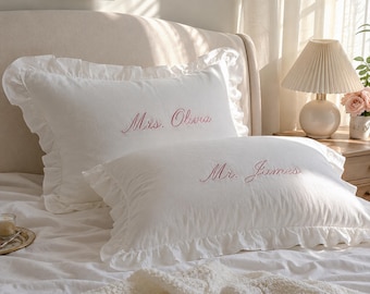 Puede incluir: Dos fundas de almohada rectangulares blancas con bordes de volantes y texto bordado en rosa. La funda superior presenta el nombre Mrs. Olivia, mientras que la inferior muestra el nombre Mr. James en una fuente cursiva.