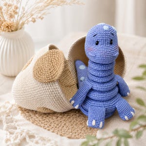 May include: A blue crocheted dinosaur toy with light blue spots and white claws, sitting inside a beige and tan crocheted egg-shaped container. The dinosaur has embroidered black eyes and pink cheeks, crafted with a textured yarn stitch pattern.