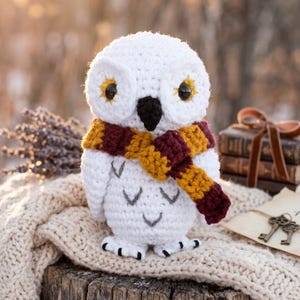 May include: A white crocheted owl plush toy featuring large black eyes with yellow accents, a black beak, and grey V-shaped feather details on its body. The owl wears a thick, striped scarf in alternating bands of dark red and mustard yellow.