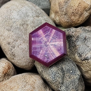 Può includere: Una gemma esagonale color magenta intenso caratterizzata da un distinto motivo a stella a sei punte che si irradia dal centro. La pietra presenta una superficie liscia e lucida con inclusioni interne che riflettono la luce.