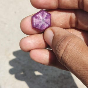 May include: A hexagonal-cut ruby gemstone featuring a distinct six-rayed star pattern across its deep magenta surface. The stone displays a translucent quality with visible internal inclusions and a polished finish, showcasing a geometric shape.