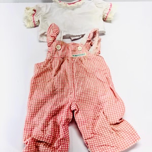 May include: A white short-sleeved shirt with ruffled lace trim on the sleeves and collar, paired with pink and white gingham overalls featuring two front buttons and a small green rectangular label on the chest area.