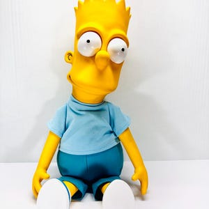 May include: A yellow Bart Simpson character doll with spiky hair and large white eyes. The figure wears a light blue short-sleeved shirt, dark blue shorts, and white shoes. The doll is in a seated position with its arms resting at its sides.