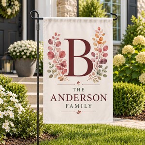 Könnte beinhalten: Eine rechteckige Gartenflagge mit einem großen dunkelroten Buchstaben B, der zwischen zwei botanischen Zweigen im Aquarellstil mit herbstlich gefärbten Blättern zentriert ist. Unter dem Buchstaben steht der Text THE ANDERSON FAMILY in Dunkelrot und Grau.