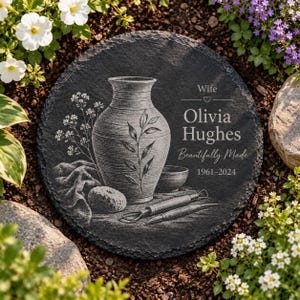 May include: A round black slate memorial plaque featuring an engraved illustration of a pottery vase, a small bowl, and sculpting tools. The text reads: Wife, Olivia Hughes, Beautifully Made, 1961-2024. The plaque has a rough, textured edge.