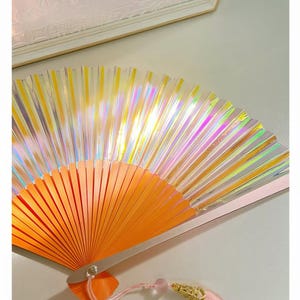 May include: A handheld folding fan featuring a bright orange frame and iridescent, holographic pleated fabric that reflects light in various colours. A light pink tassel with a gold-toned metal cap and a small bead is attached to the base.