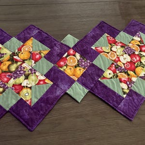 Op de afbeelding: Een gewatteerde tafelloper met een stervormig patroon en een kleurrijke fruitprint met sinaasappels, appels en druiven. Het ontwerp is omrand met effen paarse stof en lichtgroene driehoekige accenten, wat zorgt voor een geometrische indeling.