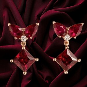 Könnte beinhalten: Ein Paar Ohrringe aus Roségold mit zwei birnenförmigen, tiefroten Rubinen an der Oberseite, akzentuiert durch einen kleinen runden weißen Diamanten, und einem hängenden quadratischen, tiefroten Rubin, gefasst in einen polierten Metallrahmen.