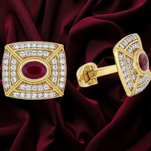 May include: A pair of gold cufflinks featuring a rectangular face with a central oval-cut deep red gemstone surrounded by rows of small clear white stones. The metal frame has a textured border and a hinged toggle mechanism on the back.