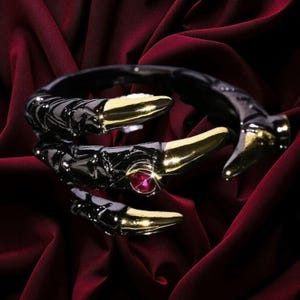 May include: A wrap-around ring featuring a black textured band shaped like a claw with polished gold-tone tips. The design includes a small, round, faceted red gemstone set into the side of the claw, creating a striking contrast against the dark metal finish.