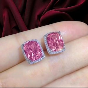 Puede incluir: Un par de pendientes de botón con piedras centrales rosas rectangulares rodeadas por un halo de pequeñas piedras claras y brillantes. Los pendientes tienen un engaste metálico y se muestran sobre un fondo de tela oscura drapeada.