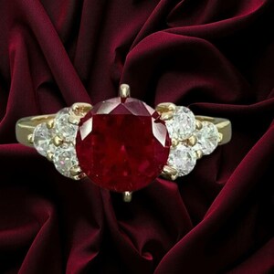Könnte beinhalten: Ein Goldring mit einem großen, runden, tiefroten Mittelstein, der von Krappen gehalten wird. Der Mittelstein ist auf jeder Seite von drei kleineren, klaren, rund geschliffenen Steinen in einer Gruppe flankiert, gefasst in eine glatte Goldschiene.