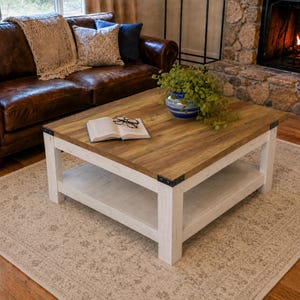 May include: A square coffee table with a natural wood grain top and a distressed white base. The table features a lower shelf, black metal corner brackets, and a sturdy four-legged frame. An open book and a potted green plant sit on the surface.