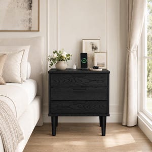 May include: A black wooden nightstand with three drawers, featuring a visible wood grain texture and four tapered legs. On top sits a small ceramic vase with white flowers, a black wireless charging station, and a small circular tray.
