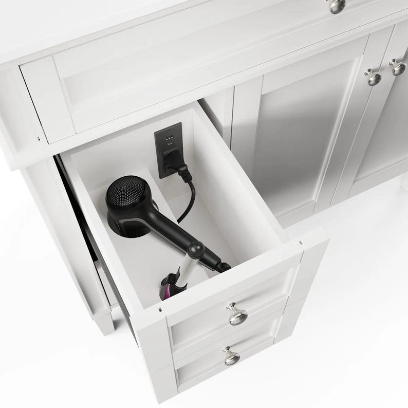 May include: A white bathroom vanity cabinet featuring an open top drawer with a built-in electrical outlet. A black hair dryer is plugged into the outlet and rests inside the drawer, which also includes a second closed drawer below it.