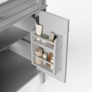 May include: A light gray bathroom vanity cabinet with an open door featuring an attached two-tier storage rack. The rack holds various personal care items including tubes, a rolled beige towel, a small wooden comb, and small bottles.