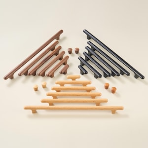 Puede incluir: Una colección de herrajes de madera para muebles que incluye tiradores en forma de T de varias longitudes y pequeños pomos redondos. El conjunto presenta tres acabados distintos: nogal marrón oscuro, negro mate y roble natural claro, dispuestos en tres grupos.