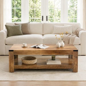 May include: A rectangular wooden coffee table with a natural grain finish and black metal corner accents. The table features a lower shelf holding a woven basket, a stack of books, and a small candle. Two white vases with yellow flowers sit on top.