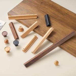 May include: A collection of cabinet hardware including various wooden and black metal drawer pulls and knobs. The assortment features long linear handles in light and dark wood tones, alongside round knobs and a single black triangular handle.