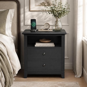 May include: A dark charcoal gray wooden nightstand featuring an open top shelf and two lower drawers with small round knobs. The nightstand holds a wireless charging stand, a small analog clock, a wooden bowl, and two stacked books.