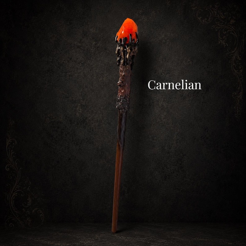 May include: A wand featuring a bright orange carnelian stone at the top, held by a dark, textured, branch-like structure that transitions into a smooth, brown, tapered handle. The word Carnelian is printed in white text to the right of the wand.
