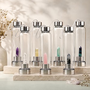 May include: Seven clear glass water bottles with silver-toned metal lids and bases. Each bottle contains a single vertical crystal point inside, featuring various colors including purple, clear, yellow, pink, light green, dark gray, and blue-green.