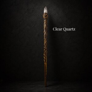 May include: A long, slender wand featuring a clear quartz crystal point at the top, secured by a textured, dark metallic setting. The shaft is brown with a winding, vine-like metallic overlay. The text Clear Quartz is visible on the right side.