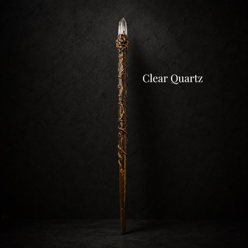May include: A long, slender wand featuring a clear quartz crystal point at the top, secured by a textured, dark metallic setting. The shaft is brown with a winding, vine-like metallic overlay. The text Clear Quartz is visible on the right side.