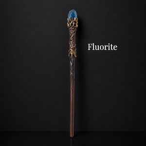 May include: A wand featuring a dark brown, twisted handle and a gold-colored metal crown at the top. A blue, translucent, rough-cut crystal is set within the crown. The word Fluorite is printed in white text to the right of the wand.