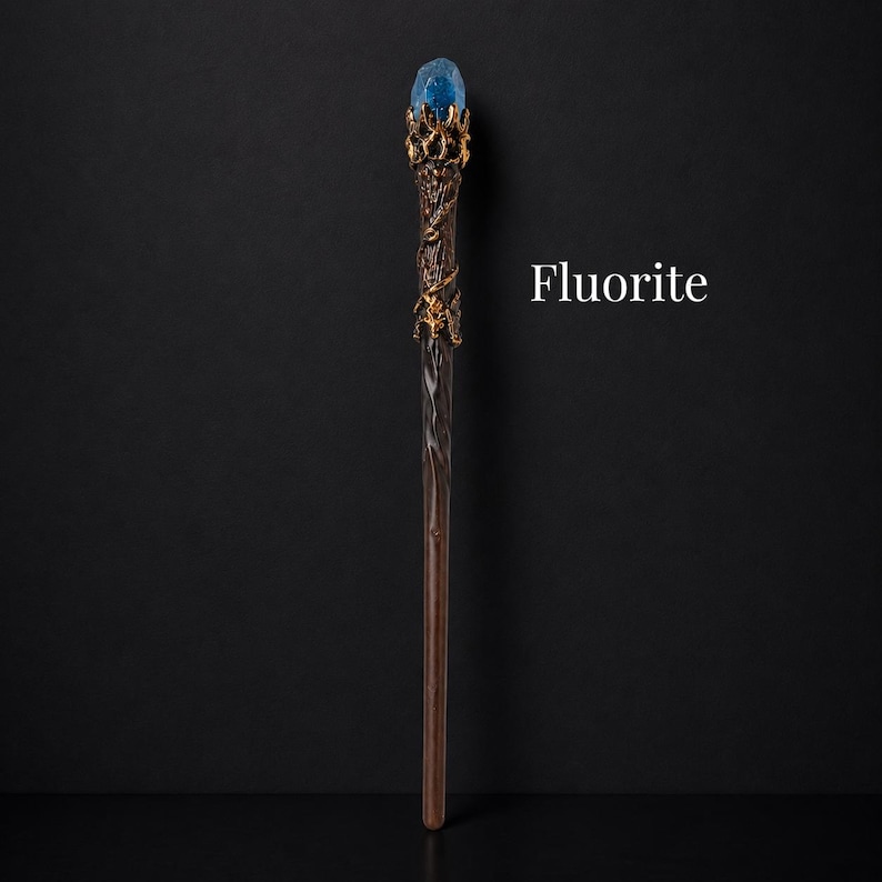 May include: A wand featuring a dark brown, twisted handle and a gold-colored metal crown at the top. A blue, translucent, rough-cut crystal is set within the crown. The word Fluorite is printed in white text to the right of the wand.