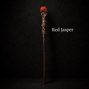 May include: A wand featuring a dark brown wooden shaft and a textured, metallic bronze-colored handle topped with a rough, vibrant red jasper stone. The text Red Jasper is printed in white to the right of the wand against a dark background.