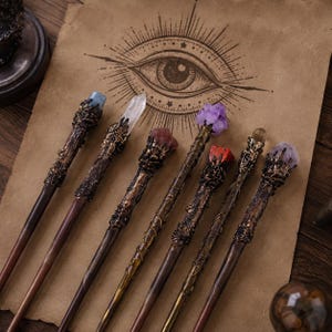 May include: Seven wands with dark brown, textured, branch-like handles and metallic, claw-like settings holding various crystals, including clear, blue, purple, and red stones, arranged on a tan paper sheet featuring a printed eye illustration.