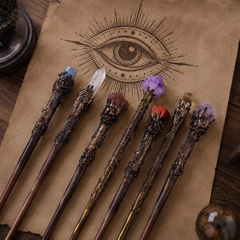 May include: Seven wands with dark brown, textured, branch-like handles and metallic, claw-like settings holding various crystals, including clear, blue, purple, and red stones, arranged on a tan paper sheet featuring a printed eye illustration.