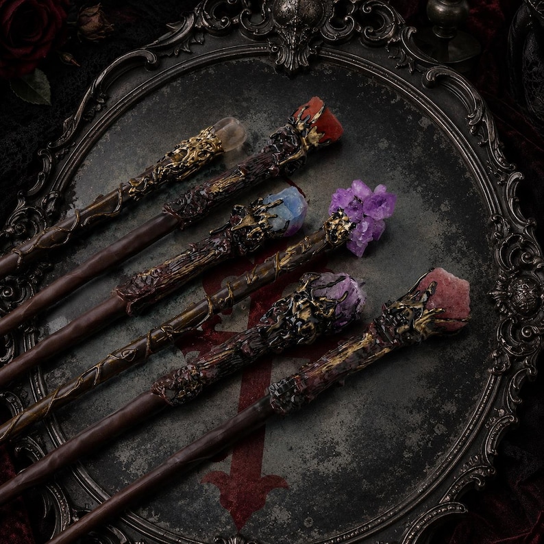 May include: Five wands with dark brown shafts and intricate gold-toned metal detailing at the handles. Each wand is topped with a unique raw crystal, including clear, blue, purple, and reddish-pink stones, arranged on a dark ornate tray.