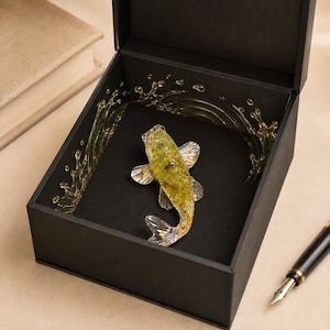 May include: A black gift box containing a translucent yellow and gold koi fish figurine. The interior of the box features a decorative gold-colored water splash design surrounding the fish. A black fountain pen rests outside the box.
